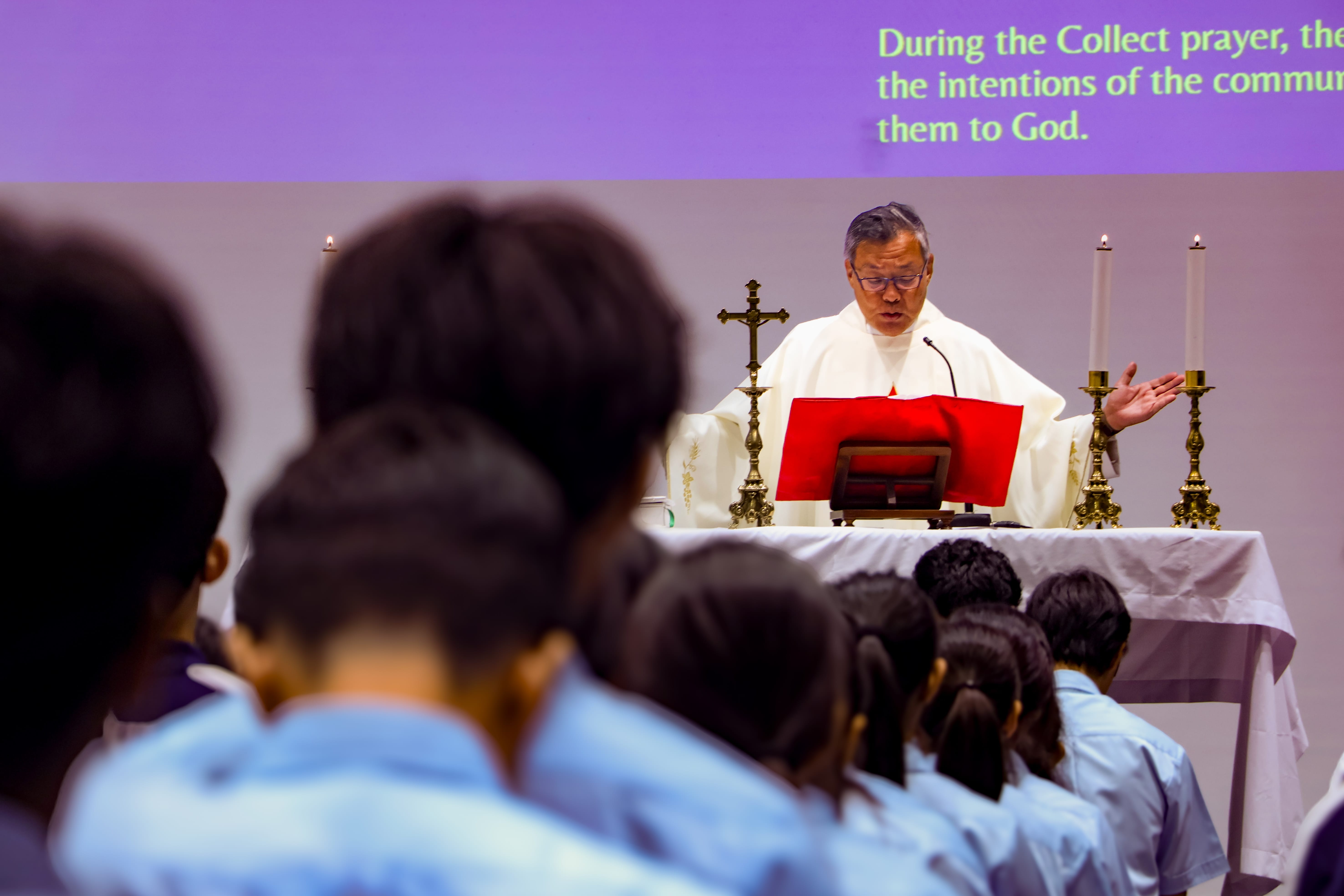 A priest in white vestments leads Mass at an altar with candles and a cross, facing seated students in blue uniforms.